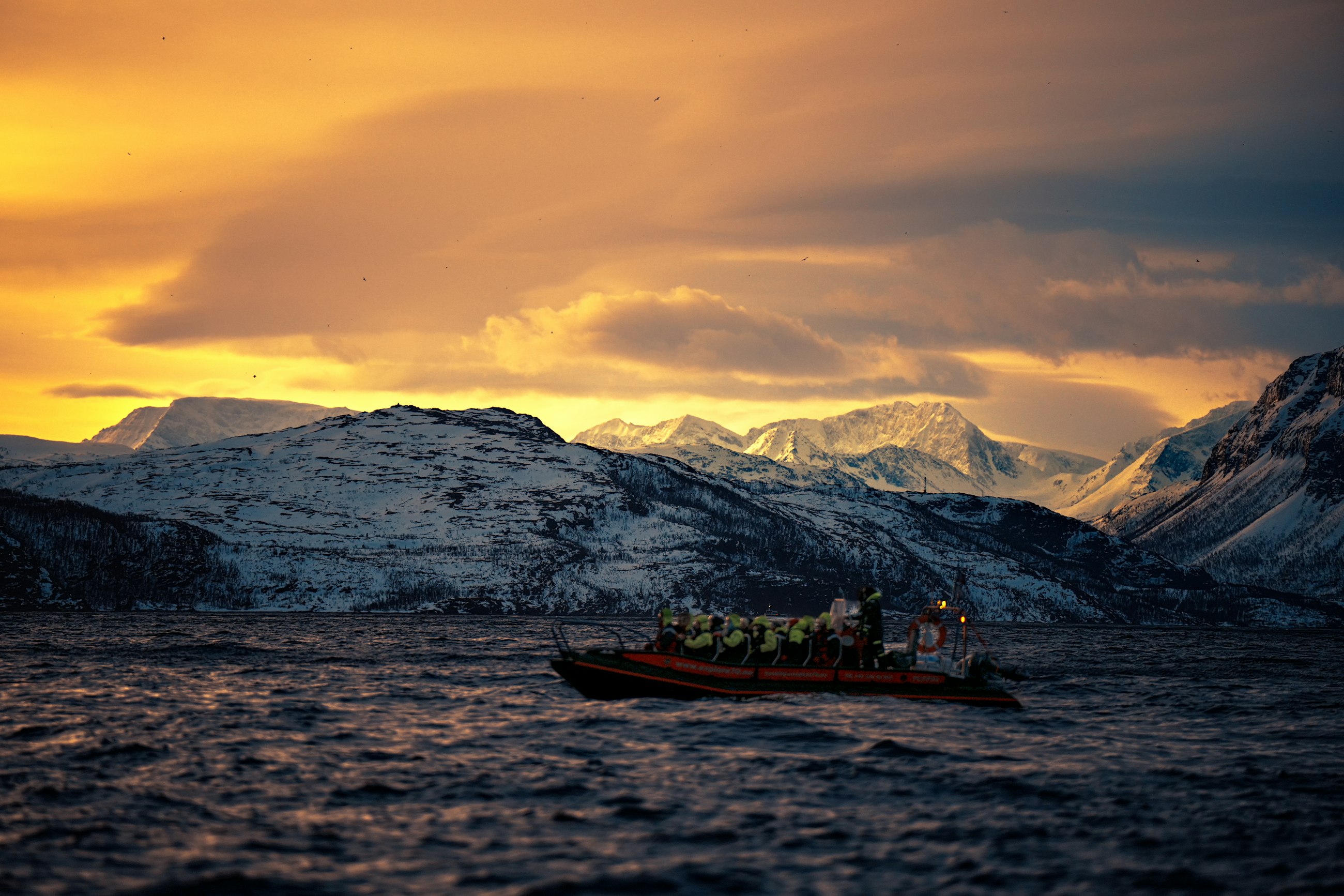 Båt med mannskap i overlevingsdrakter på fjorden utenfor Tromsø, i midnattssol med snødekte fjell bak.